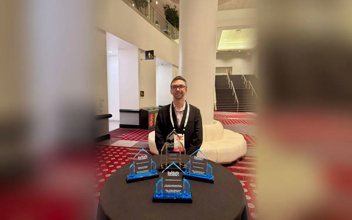 Gavin Rozzi at the 2026 NAHRO National Conference in Denver, standing behind four award trophies engraved for the New Jersey Department of Community Affairs: a NAHRO National Award of Excellence and three NAHRO National Awards of Merit.