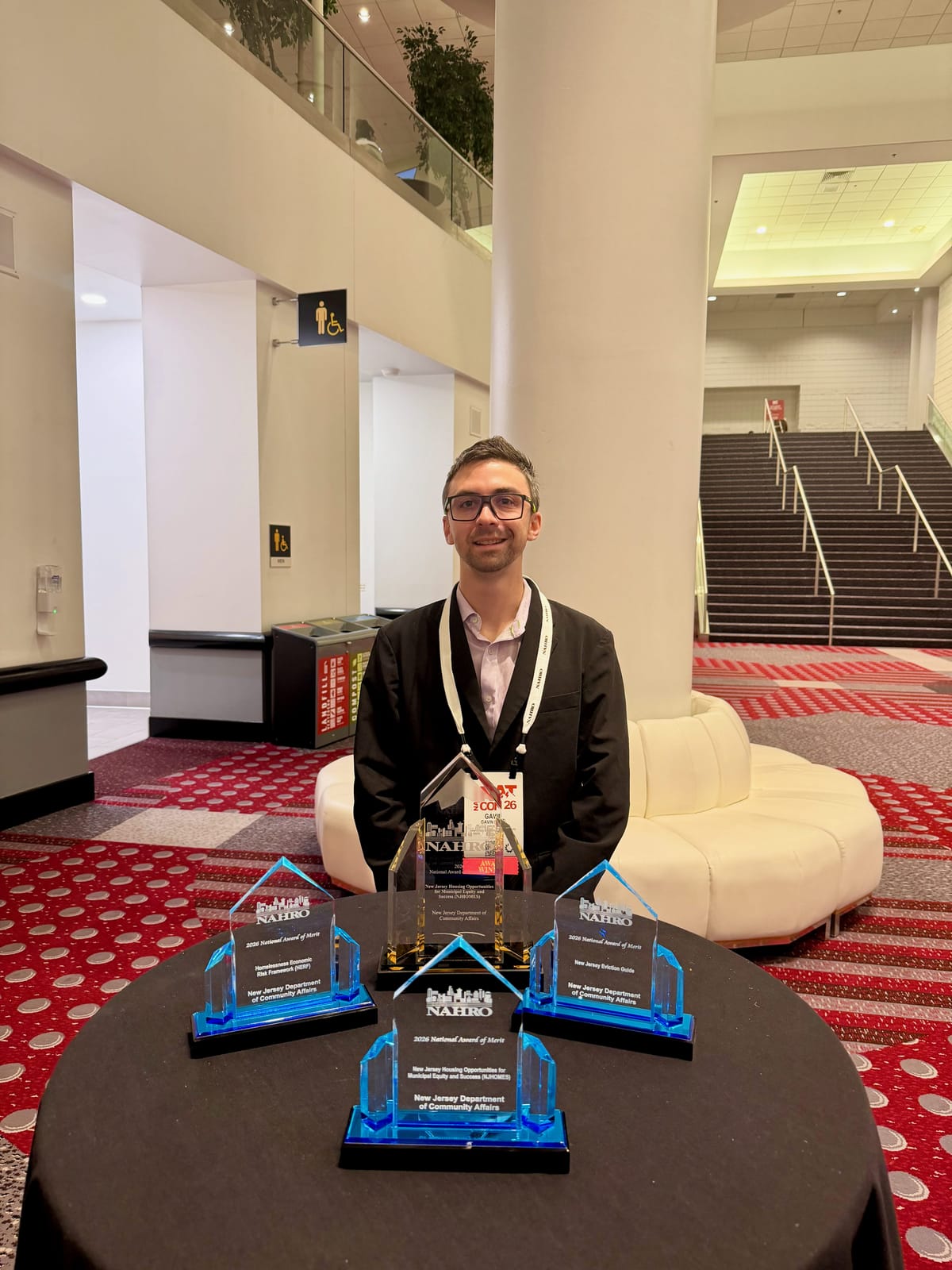Gavin Rozzi at the 2026 NAHRO National Conference in Denver, standing behind four award trophies engraved for the New Jersey Department of Community Affairs: a NAHRO National Award of Excellence and three NAHRO National Awards of Merit.